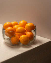 The Collective Pebbled Salad Bowl, made from clear recycled glass, holds whole oranges and sits on a beige textured surface against a plain light background.
