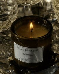 A dark green Coriandoli Candle by Flamingo Estate burns gently, its vegetable wax casting a warm glow amid glassware and a metallic surface, adding elegance to any setting.