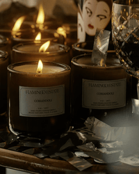 The Coriandoli Candle by Flamingo Estate, in brown glass jars, sits on a reflective surface with scattered metallic foil, a crystal glass, and a decorative cup in the background—highlighting this limited edition piece.