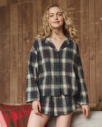 A woman with wavy blonde hair smiles while wearing the Great. Sleep’s Flannel Pajama Top in Pine Lodge Plaid, standing indoors against a wooden wall.