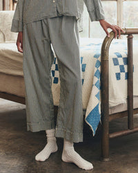A person stands by a metal bed frame wearing the Great. Sleep's Easy Sleep Pant in Black & Cream Crew Stripe and white socks. A blue and white square-patterned quilt covers the bed; the person's head is out of frame.