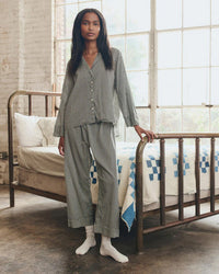 A person wearing the Great. Sleep Easy Sleep Pant in Black & Cream Crew Stripe stands by a vintage metal bed made with pillows and a white quilt with blue squares, as sunlight streams through large windows.