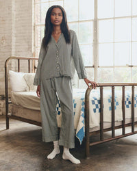 A woman stands by a vintage metal bed in a sunlit room, wearing the Great. Sleep’s Pajama Shirt in Black & Cream Crew Stripe with white socks. The bed features a white and blue quilt, while large windows fill the space with natural light.