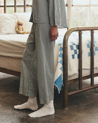 A person wearing The Easy Sleep Pant in Black & Cream Crew Stripe by the Great. Sleep stands by a brass bed with a blue and white quilt; a teddy bear sits on a pillow in the background.