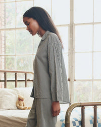 A woman wearing the Great. Sleep’s Pajama Shirt in Black & Cream Crew Stripe stands beside a brass bed with a teddy bear and quilt, bathed in sunlight from large window panes.