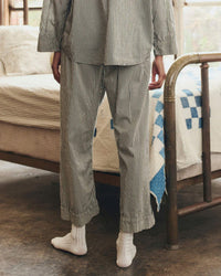 Wearing The Easy Sleep Pant in Black & Cream Crew Stripe by the Great. Sleep, a person stands near a metal-framed bed with a white and blue patchwork quilt in a cozy, sunlit room. Only their lower body is shown.