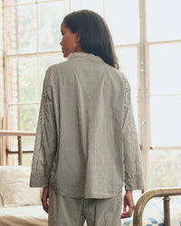 A woman with long dark hair stands indoors, her back to the camera, wearing the Great. Sleep’s Pajama Shirt in Black & Cream Crew Stripe. Natural light streams through large windows in the background.