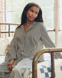 A woman with long dark hair wears the Great. Sleep's Pajama Shirt in Black & Cream Crew Stripe while sitting on a bed with a brass frame and blue-and-white quilt, surrounded by large windows letting in natural light.