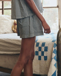 A person in the Great. Sleep’s Sleep Boxer Short in Black & Cream Crew Stripe, featuring an elastic waistband and matching top, stands by a bed with a blue and white patchwork quilt; a stuffed animal rests on the pillow behind them.