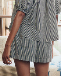 A person wears The Sleep Boxer Short in Black & Cream Crew Stripe by the Great. Sleep—100% cotton with an elastic waistband—matching a black-and-white vertically striped short-sleeve top, standing indoors with their arm relaxed by their side.
