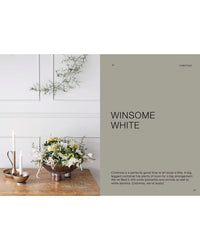 A minimalist holiday scene with white walls, florals in a bronze vessel, two glowing candles, and wall greenery. Text: “The Potted Garden - All Year Round” by Cozy Publishing—your guide to seasonal Christmas arrangements.