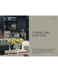 A modern patio features black planters from "The Potted Garden - All Year Round" by Cozy Publishing, with sunflowers and lush greenery in gray, brown, and yellow tones set against a glass building backdrop.