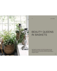 Two baskets featuring green and white seasonal arrangements from Cozy Publishing’s "The Potted Garden - All Year Round" decorate a table by the window, their fresh look described as “Beauty Queens in Baskets.”.