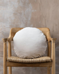The Saarde Linen/Velvet Round Pillow with Feather Insert in Clay rests on a wooden chair with a woven seat, set against a textured beige wall.