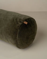 The Saarde Velvet Structured Bolster in Dark Olive, a cylindrical cushion with a small fabric tag, is shown resting on a light surface.