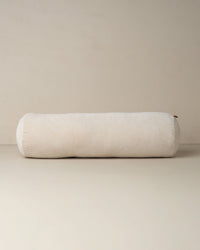 The Saarde Velvet Structured Bolster in Clay, featuring a large cylindrical shape, rests on a neutral beige surface with a matching backdrop.