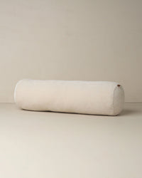 The Saarde Velvet Structured Bolster in Clay, a long cylindrical cushion, rests on a light-colored floor against a plain, light wall.