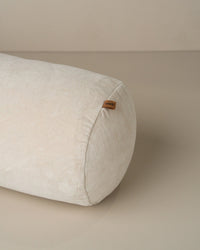 The Saarde Velvet Structured Bolster in Clay is a cylindrical off-white cushion with a small brown fabric tag on one end, displayed on a smooth, neutral surface.