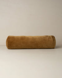 The Saarde Velvet Structured Bolster in Nutmeg, a long, cylindrical velvet cushion in rich brown, rests on a plain light surface against a neutral wall.