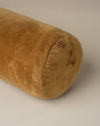 The Saarde Velvet Structured Bolster in Nutmeg, a cylindrical cotton velvet cushion with a small fabric tag on the seam, rests on a light surface.
