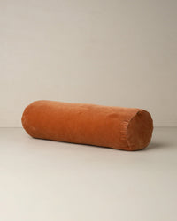 The Saarde Velvet Structured Bolster in Terracotta, featuring a cylindrical shape and soft cotton velvet, rests on a neutral light surface against a plain wall.