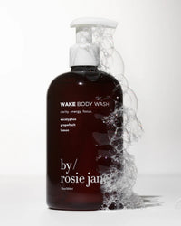 A dark amber pump bottle of Rosie Jane Wake Body Wash, covered in soap bubbles, stands against a white background. The label highlights its antioxidant-rich formula with eucalyptus, grapefruit, and lemon ingredients.
