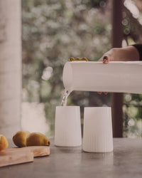 A hand pours water from a white pitcher into a Saarde Kairos Large Water Glass in Opaque White on a table with lemons and a wooden board nearby.