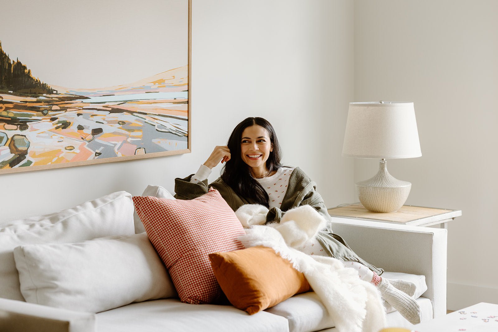 A woman sits on a white sofa with colorful pillows, smiling, next to a lamp and a landscape painting on the wall.