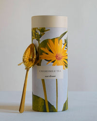 The Quiet Botanist Chamomile Tea Gift Set features a canister with a large yellow flower on the label and a gold tea infuser beside it, all set against a plain, light background.