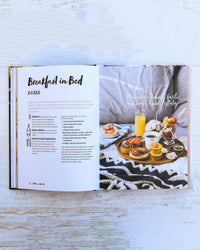 The cookbook "Platters and Boards" by Chronicle Books features a “Breakfast in Bed” recipe with a styled photo of breakfast foods, juice, and flowers arranged artfully on platters and boards over a patterned blanket.
