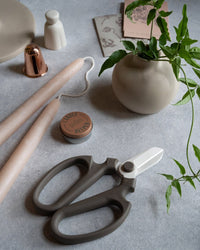 A tabletop features pink taper candles, The Floral Society’s Japanese Floral Clippers in Peat, a small potted plant, a copper candle snuffer, a round tin, and paper cards on a light gray surface.