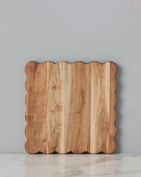 The etúHOME New Scalloped Cutting Board, Medium Square, stands upright against a light gray wall on a white marble surface.