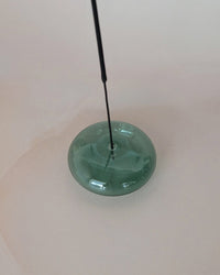 A thin black stick stands upright in the center of a round, flat, translucent teal Bubble Glass Incense Holder by BOTANICA, displayed on a light beige surface.