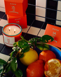 A lit ROEN La Condesa Candle made with coconut soy wax sits in a glass jar beside its boxes, fresh lemons, oranges, and leafy branches, filling the air with premium fragrance oils.