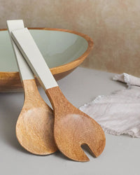 The Madras Serving Set in Taupe by Be Home US, featuring hand-carved servers with taupe handles, rests on the edge of a wooden bowl beside a light-colored napkin.