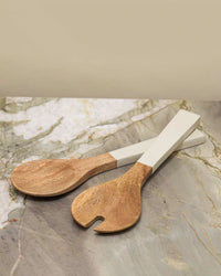 The Be Home US Madras Serving Set in Taupe features two hand-carved salad servers with white handles, crafted from sustainably sourced mango wood, resting elegantly on a marble surface.