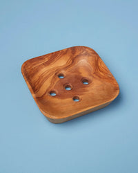 The Be Home US Olive Wood Square Soap Dish, made from sustainably harvested olive wood with five drainage holes and rounded corners, is displayed on a light blue surface.