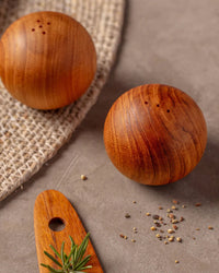 The Be Home US Teak Salt & Pepper Shaker Set, featuring two artisan-crafted round pieces, rests on a textured surface with scattered pepper and a wooden utensil garnished with rosemary nearby.