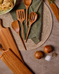 Be Home US Teak Salt & Pepper Shaker Set, wooden kitchen utensils, cutting boards, and a bowl of pasta are displayed on a woven placemat atop a gray countertop.