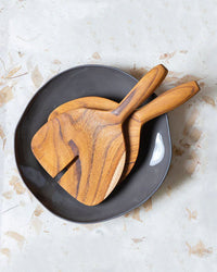 The Be Home US Teak Wide Serving Set is displayed inside a dark gray, uniquely shaped bowl on a light, textured surface.
