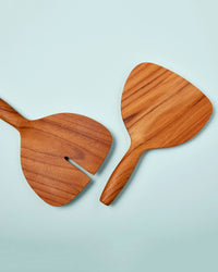 The Be Home US Teak Wide Serving Set, featuring two handcrafted wooden servers with wide, flat blades—one slotted for easy gripping—is displayed on a light blue background, combining style with elegant functionality.