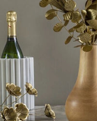 A bottle of champagne rests in the Be Home US Waterloo Marble Bottle Holder, displayed beside a wooden vase with gold metal flowers from the elegant Waterloo collection.