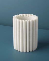 The Be Home US Waterloo Marble Bottle Holder, featuring a white cylindrical ribbed design with a fluted edge, is displayed on a blue surface against a blue background.