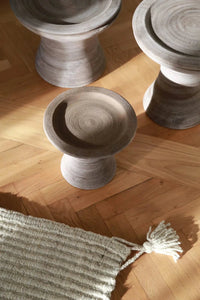 Three Bergs Potter Celeste 11 inch Pedestal Planters in Raw Grey sit on a parquet floor beside a cream tasseled rug, serving as stylish stands for plants or handcrafted pottery.