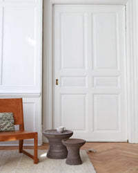 A wooden chair with a patterned pillow sits beside two round wooden side tables, one topped with a cup and the Bergs Potter Celeste 11 inch Pedestal Planter in Raw Grey, set before white paneled doors and white walls.