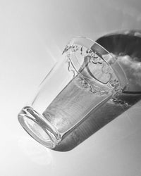 The Kobenhavner Glass Vase in Clear by Bergs Potter lies on its side on a white surface, casting a shadow and light reflections that evoke the timeless elegance of royal Danish pottery.