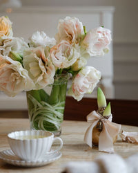 A Kobenhavner Glass Vase in Clear by Bergs Potter holds light pink flowers beside a teacup, while a ribbon-tied bulb with green shoots adds Danish pottery charm to the table.