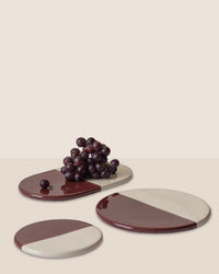 A cluster of red grapes rests on Bergs Potters Nuna Glazed 7.9 inch Tray in Sandstone/Deep Burgundy, a food-safe stoneware oval tray, alongside two matching round trays and Nuna coasters.