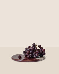 A bunch of dark purple grapes sits on the Bergs Potter Nuna Glazed 7.9 inch Tray in Sandstone/Deep Burgundy, set against a plain beige background.