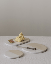 Three round marble serving boards with a butter slab, half a fig, and a silver knife sit on a white textured tablecloth with the Bergs Potter Nuna Glazed 7.9 inch Tray in Sandstone/Mineral White against a plain background.